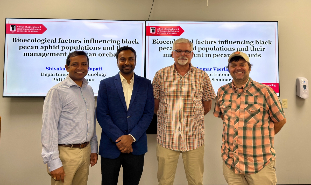 Four individuals stand in front of two display screens showing a UGA-branded presentation slide titled “Bioecological factors influencing black pecan aphid populations and their management in pecan orchards”; the group faces the camera in an indoor classroom or meeting space with neutral walls and mounted screens behind them.