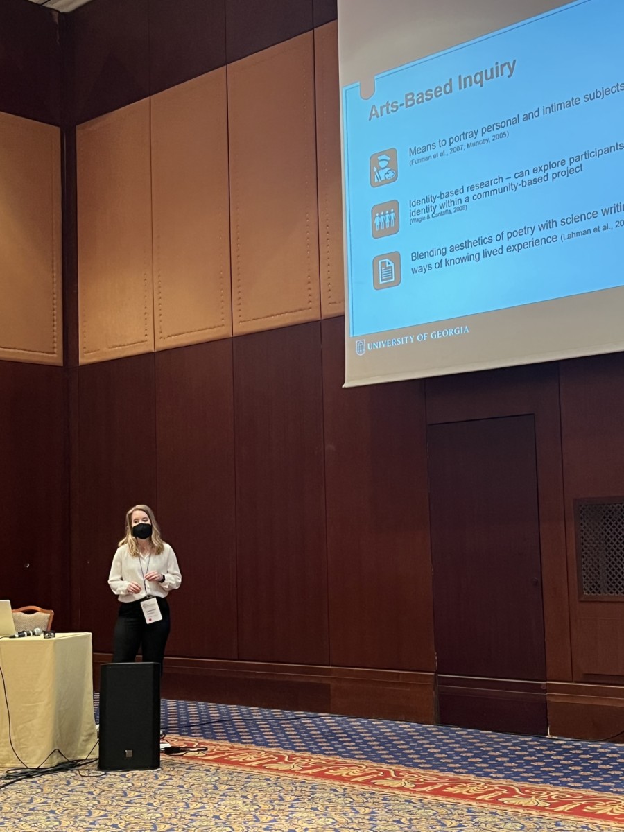 A young Caucasian woman in business casual dress presents her research on a screen